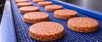 Plant-based burger patties moving on a blue food-grade conveyor belt in a food processing line for packaging meat alternatives.